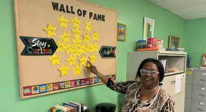 Linda Smith stands in front of a bulletin board with yellow paper stars. The stars have the names of graduates taught by Smith written on them.